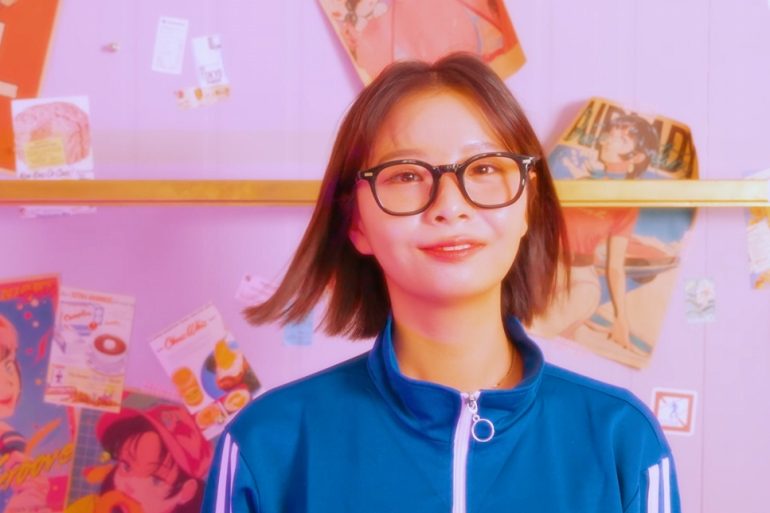 MiJoo is standing in front of a wall wearing glasses and a track suit top. Behind her on the wall are stickers, posters featuring anime girls, overalls, and a mic cord.