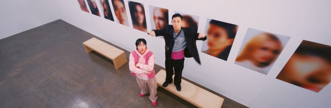 Jambino and Loco are in an art gallery. Jambino is standing with arms crossed, while Loco is standing on a bench