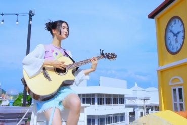 Gyubin is sitting on a small bench on a rooftop playing her guitar
