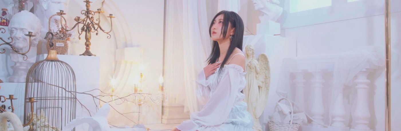 Singer CLAVE is kneeling in a white room in a white dress. Various objects surround her like statues, a bird cage, lamps, and more
