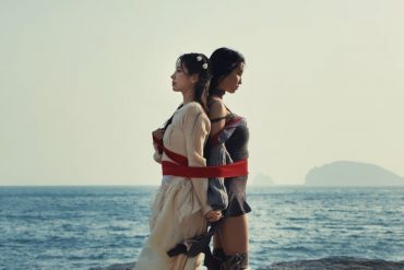 TWICE members Dahyun and Momo are standing back to back while tied together by a large ribbon. Behind them is the ocean