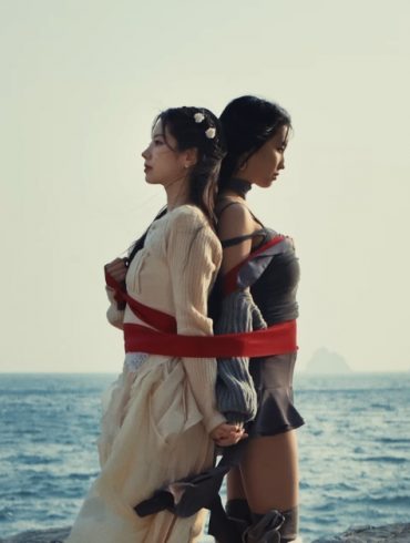 TWICE members Dahyun and Momo are standing back to back while tied together by a large ribbon. Behind them is the ocean