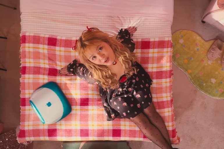 SORN sits on a bed in a very pink room. There's aa variety of items around the room like an iMAC, turntable, slippers, a Tube TV, and more.