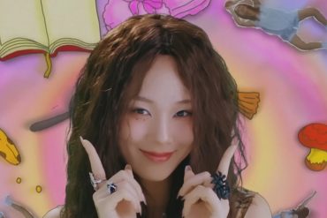 Korean singer BIBI is looking at the camera and pointing her fingers up near her face. Behind her are cartoon like items (mouse, book, broom, etc.)