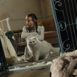 A reflection in the mirror shows YooA sitting on the floor of a messy room with a white cat