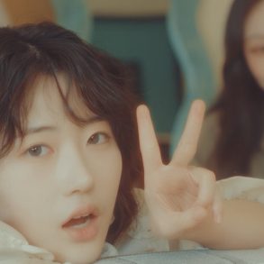 2 members of (JOY)ON are in a room. One is in focus and peering over a couch giving a peace sign. The other is in the background blurry looking at the camera.