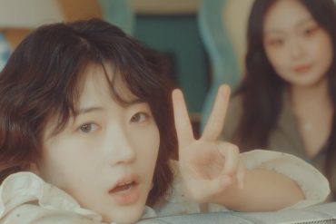 2 members of (JOY)ON are in a room. One is in focus and peering over a couch giving a peace sign. The other is in the background blurry looking at the camera.