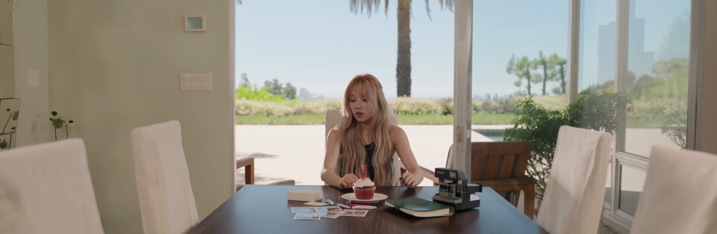 Yuqi is sitting at a table looking at a small box in front of her. Also on the table is a cupcake with a candle, a Polaroid camera, pictures, and a notebook