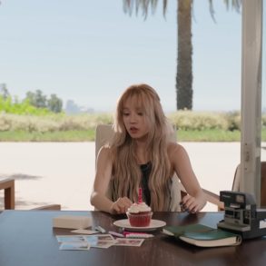 Yuqi is sitting at a table looking at a small box in front of her. Also on the table is a cupcake with a candle, a Polaroid camera, pictures, and a notebook