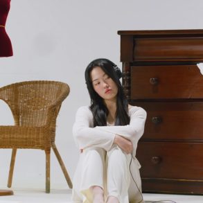 Jiwoo is sitting on the floor with her arms wrapped around her knees. There are various objects around her like a mannequin, mirror, chair, dresser, and more.
