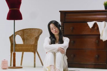 Jiwoo is sitting on the floor with her arms wrapped around her knees. There are various objects around her like a mannequin, mirror, chair, dresser, and more.