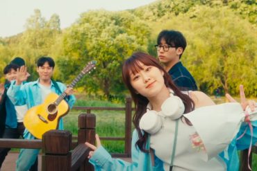 Woo Yerin is giving peace signs while standing on a wooden walkway. Behind her are 3 male band members