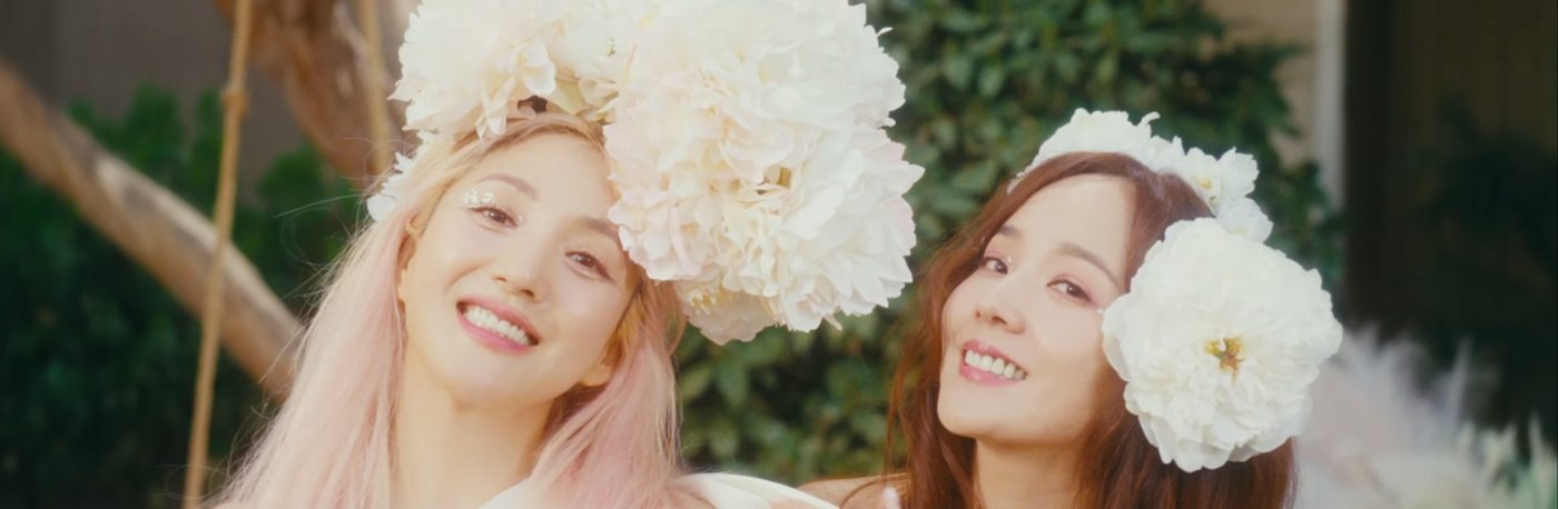 Bada and Eugene are smiling and looking at the camera. Both are wearing large flowers on their head
