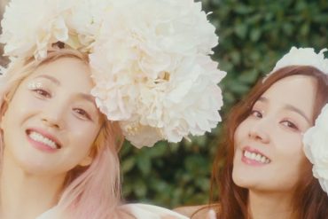 Bada and Eugene are smiling and looking at the camera. Both are wearing large flowers on their head