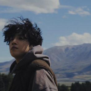 Wooseok is wearing a hoodie while looking at something off camera. There are trees and a mountain range behind him