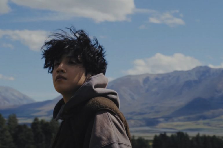 Wooseok is wearing a hoodie while looking at something off camera. There are trees and a mountain range behind him