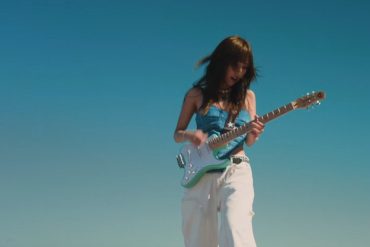 Korean singer and guitarist, MEMI, is playing a guitar with a clear, blue sky behind her