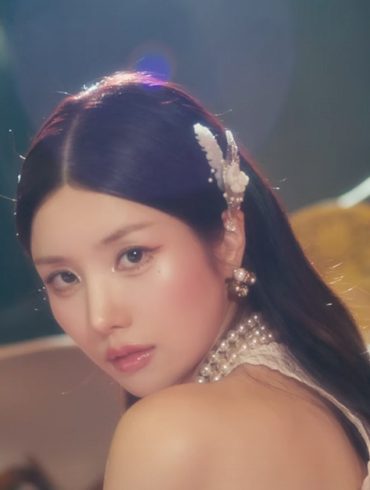 Kwon EunBi is looking back at the camera. Behind her are a bunch of chairs thrown in a wide pile