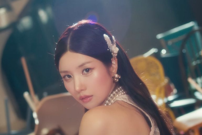 Kwon EunBi is looking back at the camera. Behind her are a bunch of chairs thrown in a wide pile