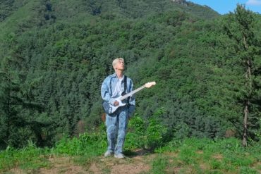 K-Pop artist BOUN stands at the edge of a cliff holding a guitar and looking up at the sky. There are mountains behind him