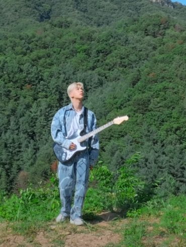 K-Pop artist BOUN stands at the edge of a cliff holding a guitar and looking up at the sky. There are mountains behind him